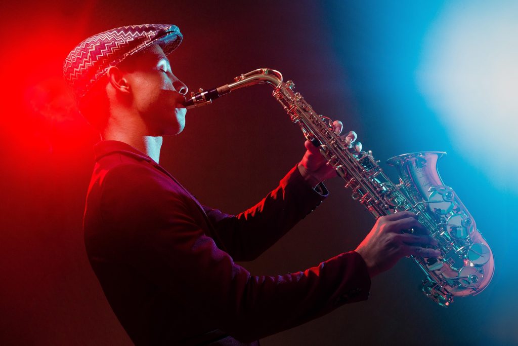 A person wearing a dark jacket and a striped cap play the saxophone in front of a red and blue light.