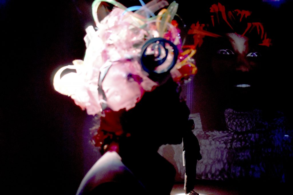 Person wearing headwear made from spirals of upcycled plastic, surrounded by the film DEMO DAY projected in 360 degrees. If possible, please load remote images to view this photo.